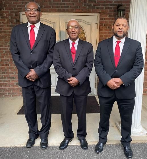 Three men in black suits with red ties standing on a front porch