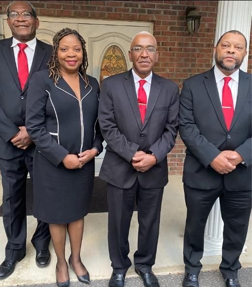 Four people in formal black suits with red ties standing together outside a brick building.