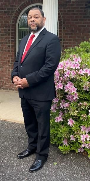 Man in a black suit with a red tie standing beside pink flowers outside a brick building