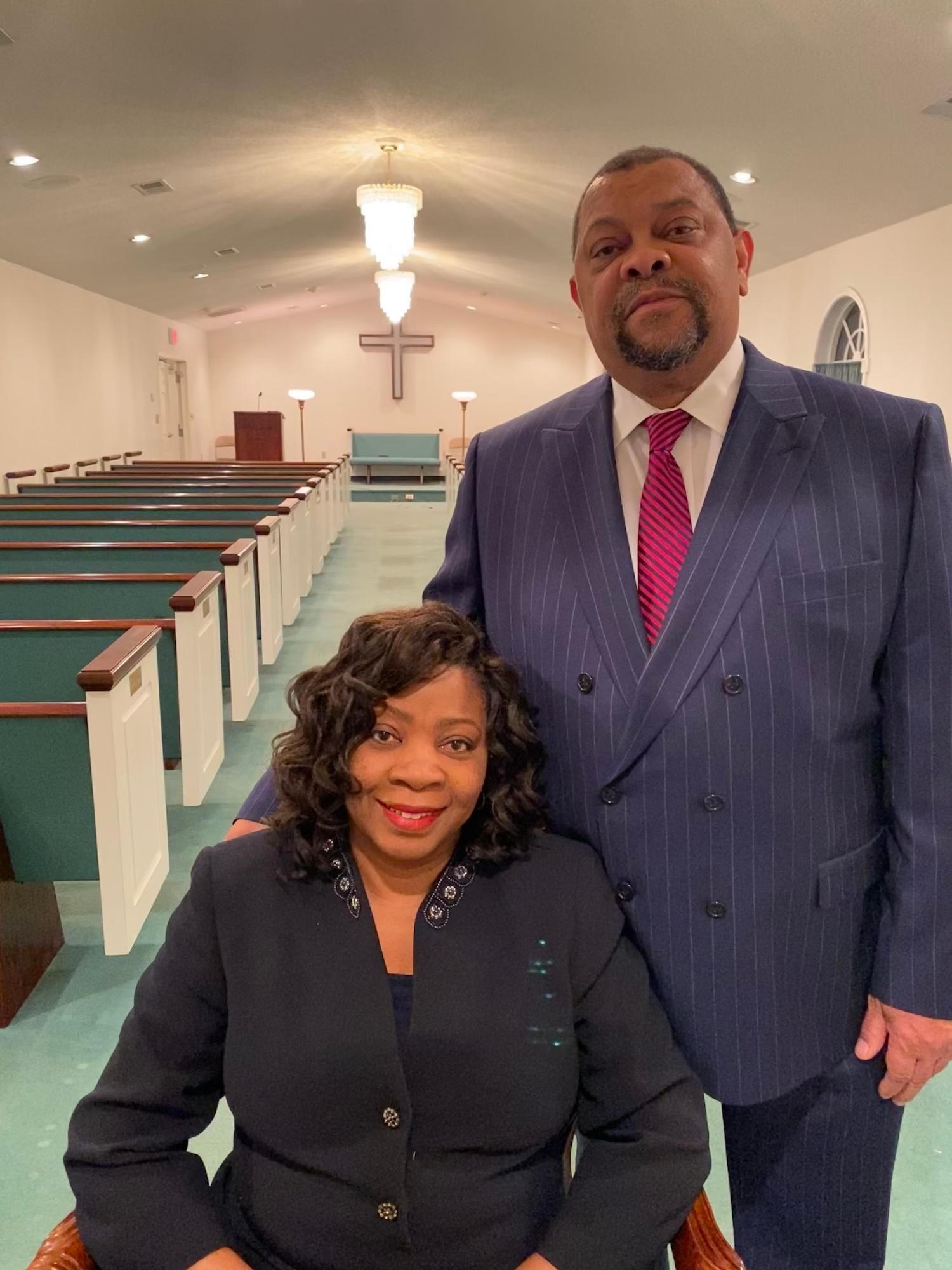 Two people posing in a church sanctuary, with pews and a cross in the background.