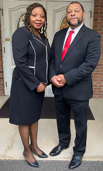 Two people in business attire standing together outdoors, posing for a formal photo.