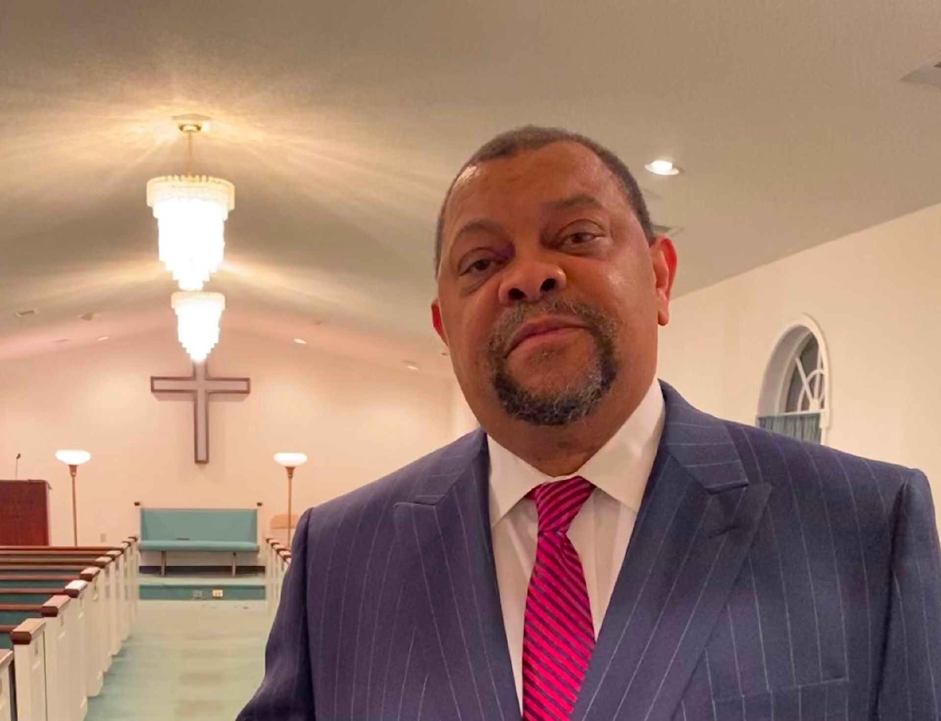 Man in a blue suit and pink tie stands in a church aisle with a cross at the altar behind him.