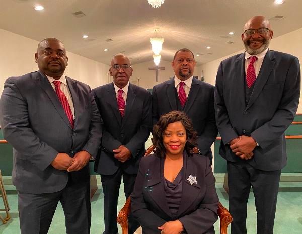 Five formally dressed people pose indoors, four standing behind one seated in front, with red ties and a teal carpet.