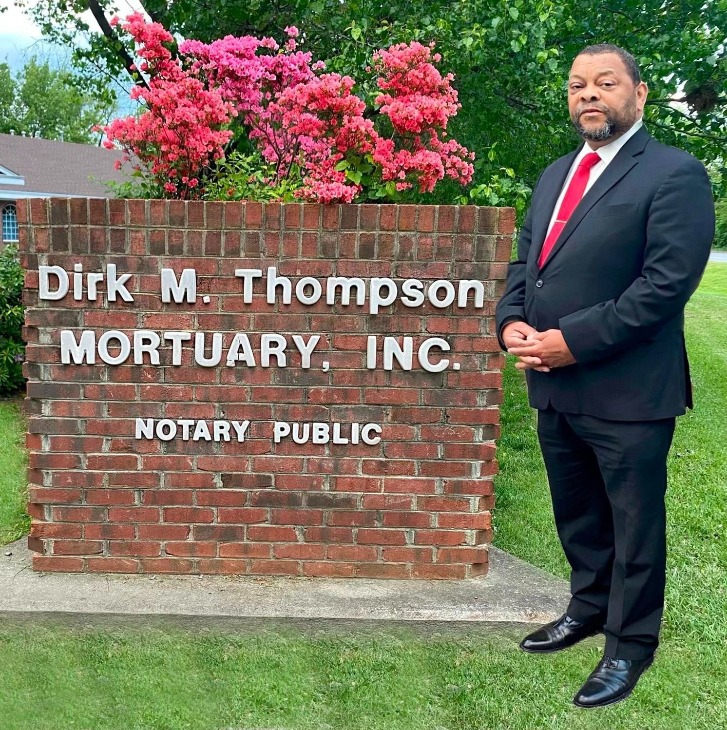 Man in dark suit beside brick sign reading “Dirk M. Thompson Mortuary, Inc.” with pink flowers behind.