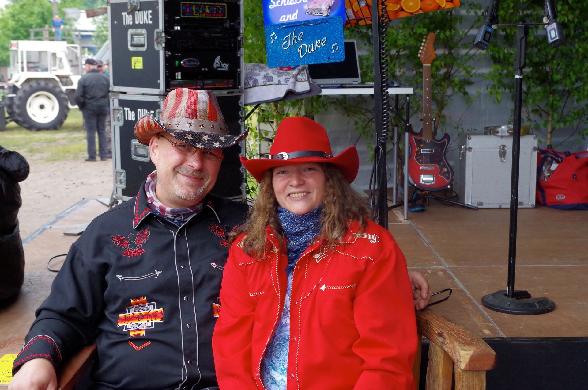 Ein Paar in Cowboyhüten und -kleidung auf der Bühne, lächelnd, im Freien, im Hintergrund Ausrüstung und ein Traktor.