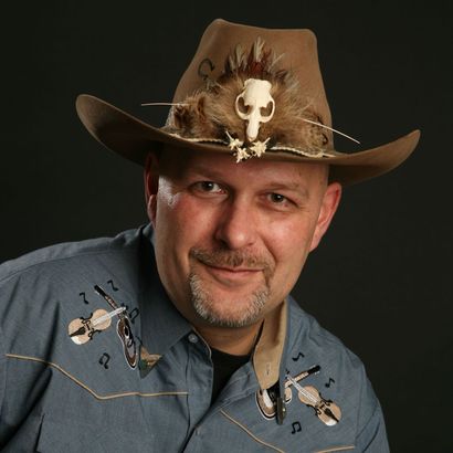 Mann mit Cowboyhut mit Totenkopfverzierung, der ein blaues Hemd mit Musikinstrumentenstickerei trägt, lächelt.