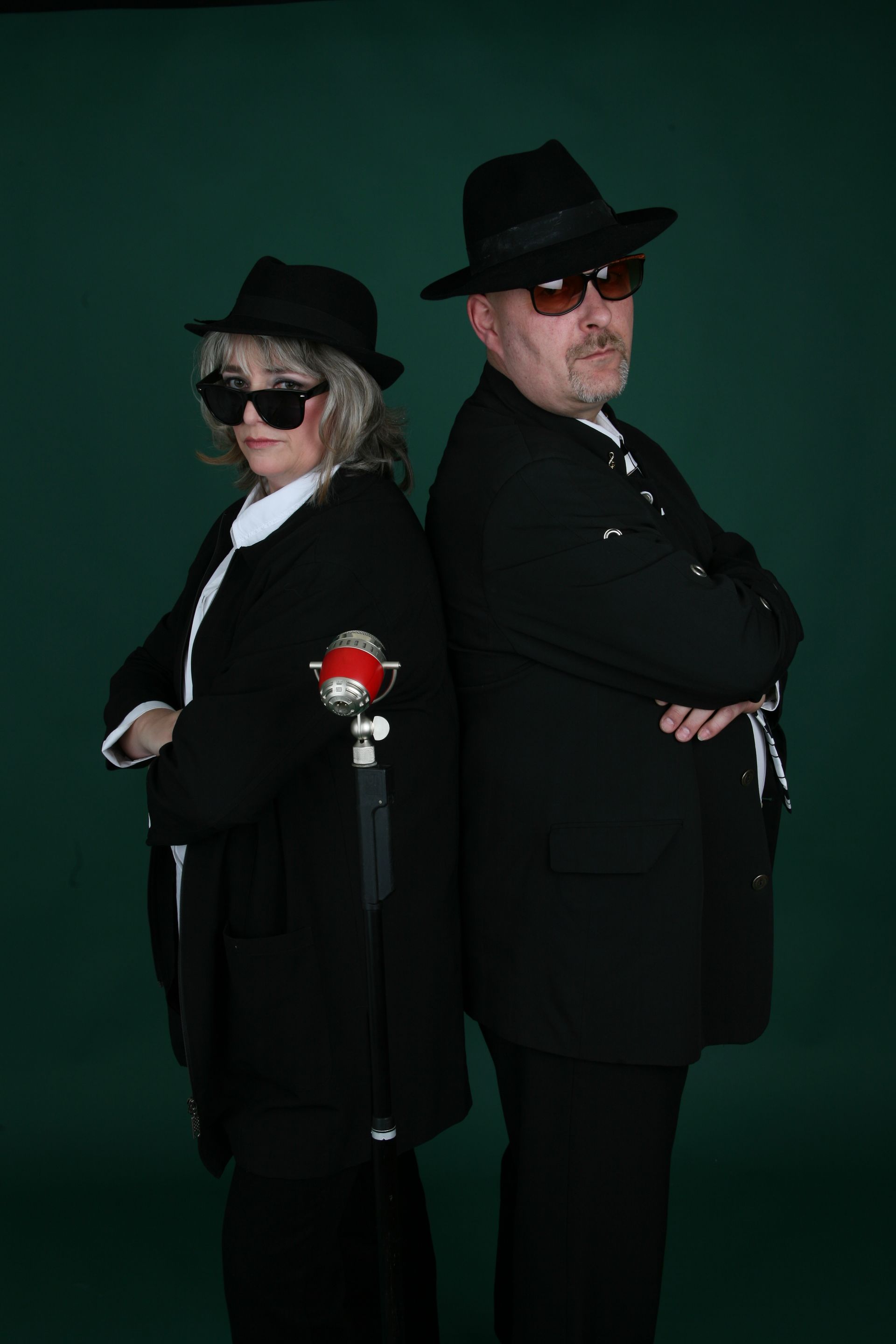 Zwei Personen, verkleidet als Blues Brothers in schwarzen Anzügen, Hüten und Sonnenbrillen, stehen Rücken an Rücken vor einem grünen Hintergrund.