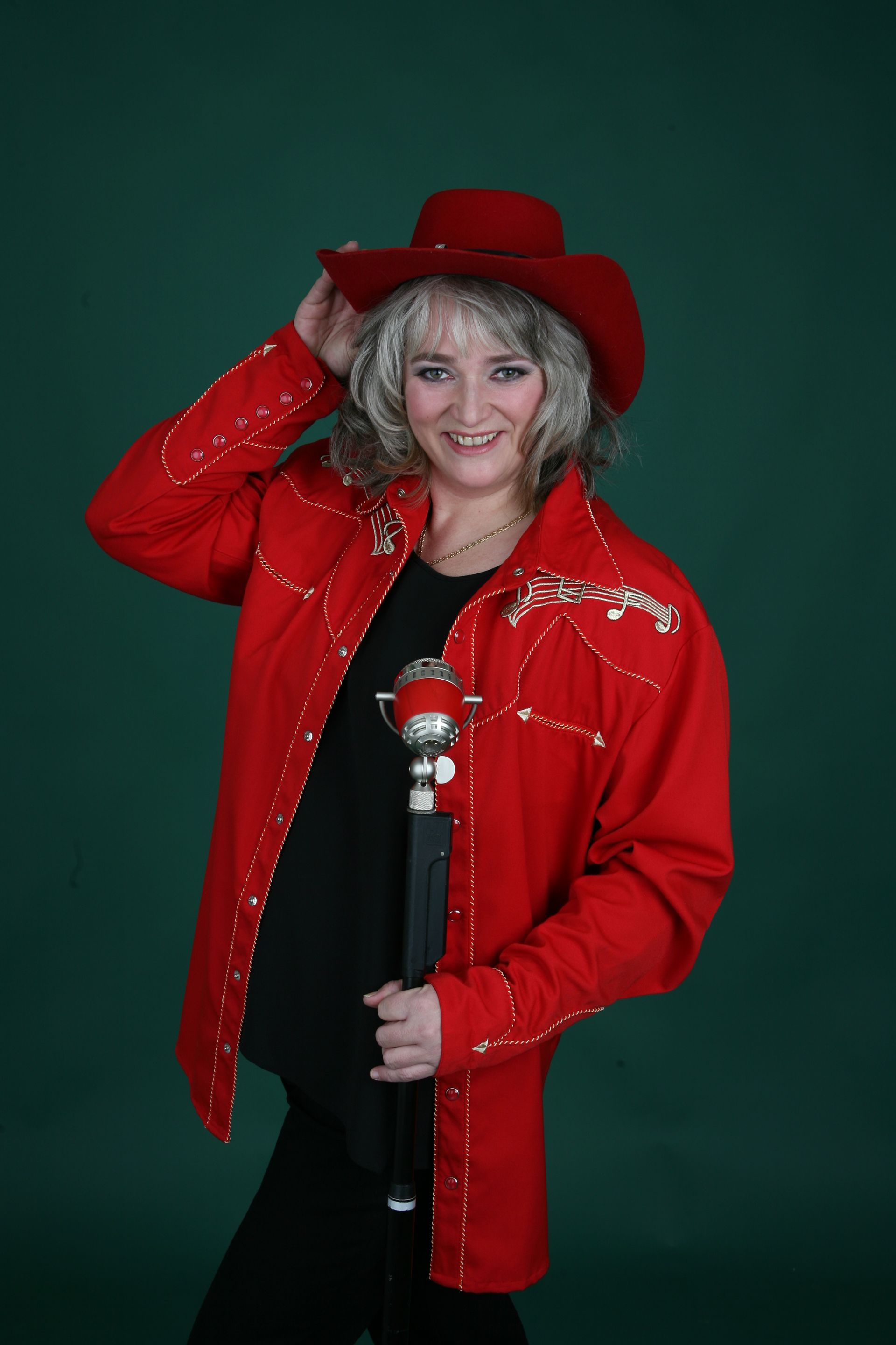 Frau mit rotem Cowboyhut und -jacke, die ein Mikrofon hält, lächelt, grüner Hintergrund.