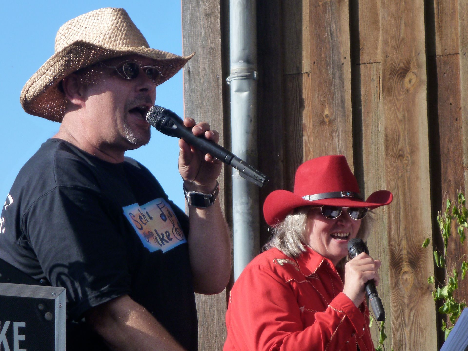 Zwei Personen singen im Freien in Mikrofone, eine trägt einen Strohhut und ein schwarzes Hemd, die andere einen roten Cowboyhut und eine rote Cowboyjacke.