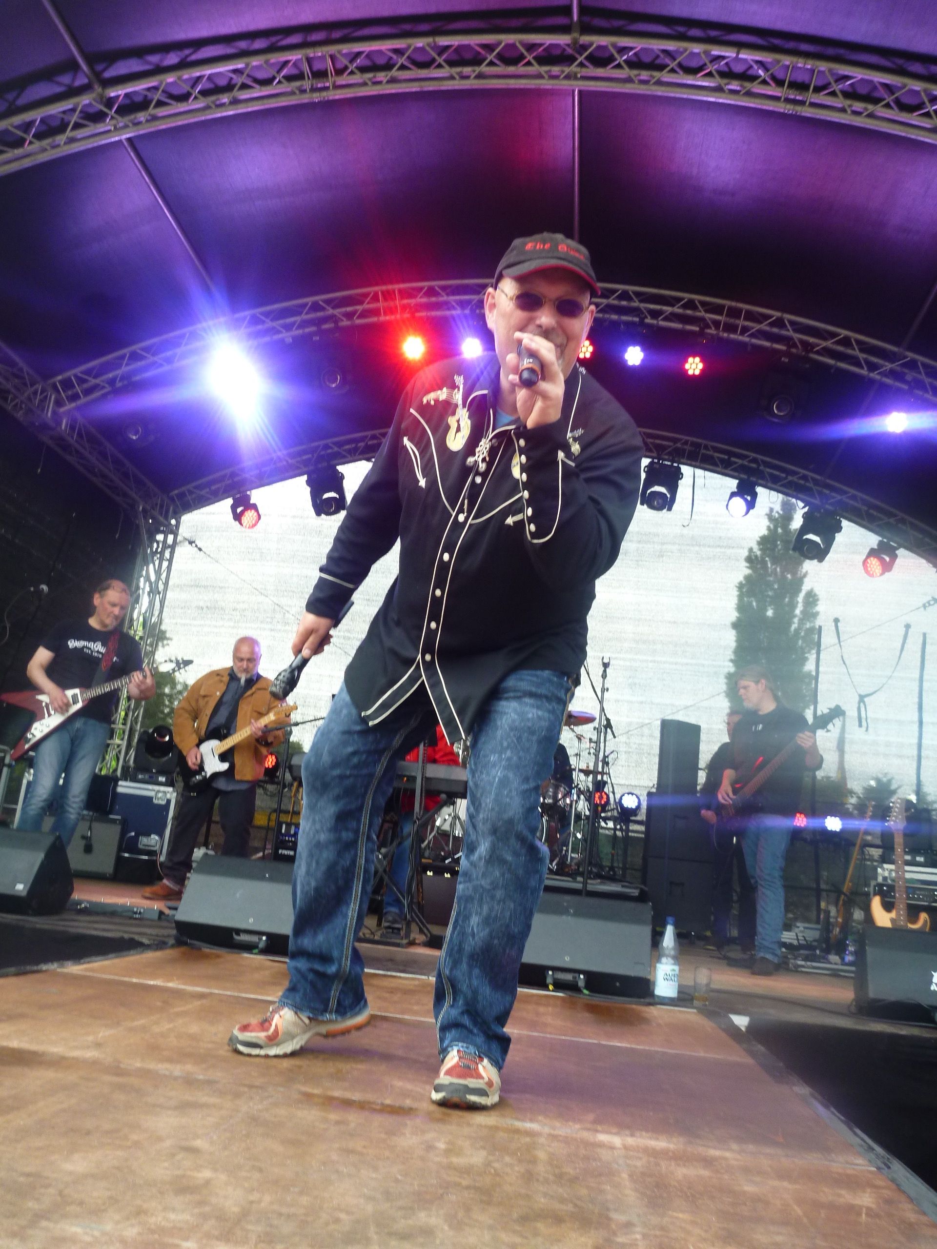Ein Mann singt auf der Bühne; er trägt ein schwarzes, verziertes Hemd, Jeans und eine Baseballkappe; im Hintergrund ist eine Band zu sehen.