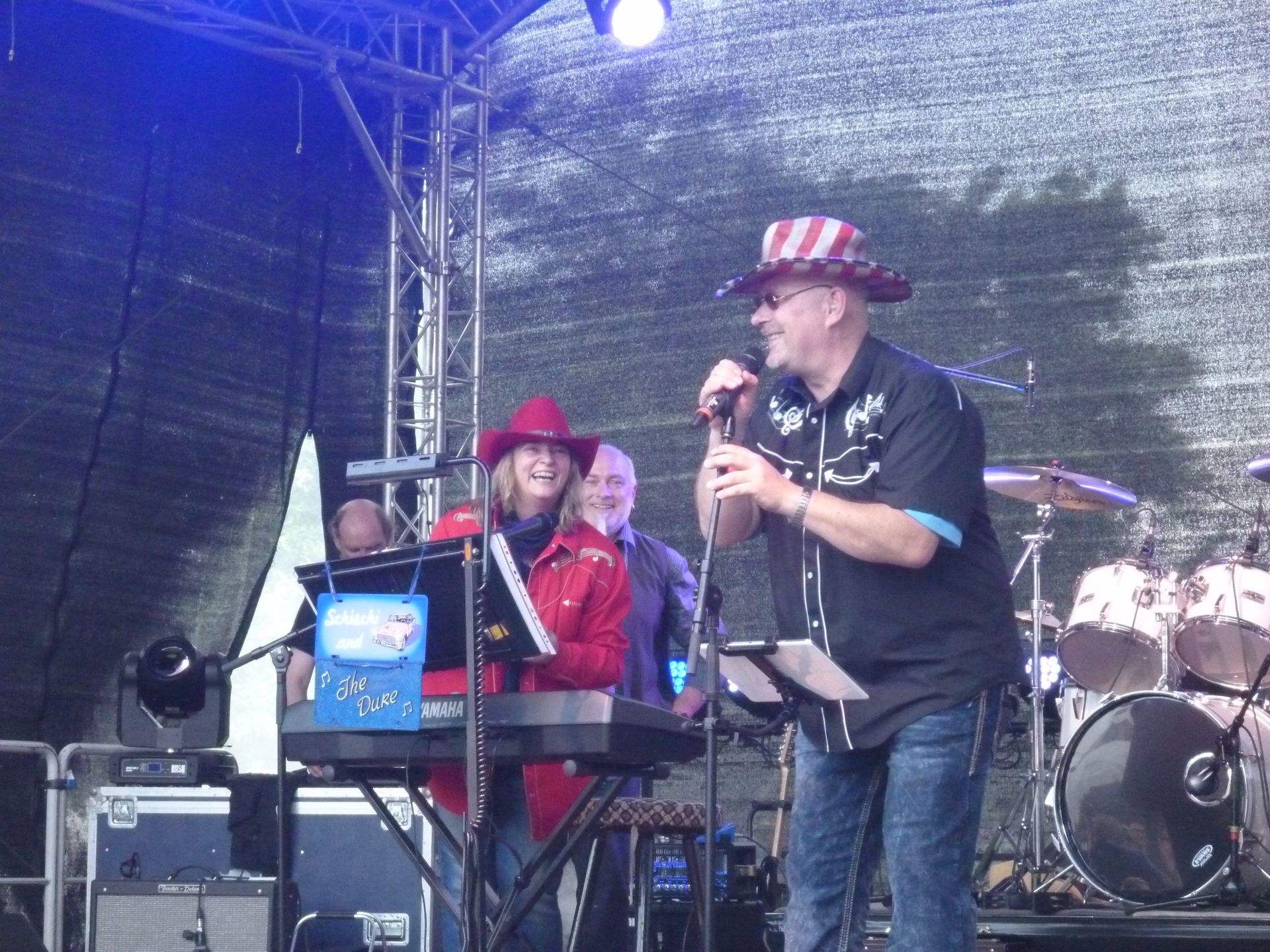 Die Band tritt auf der Bühne auf, der Sänger mit Mikrofon trägt einen Onkel-Sam-Hut, der Keyboarder eine rote Jacke.