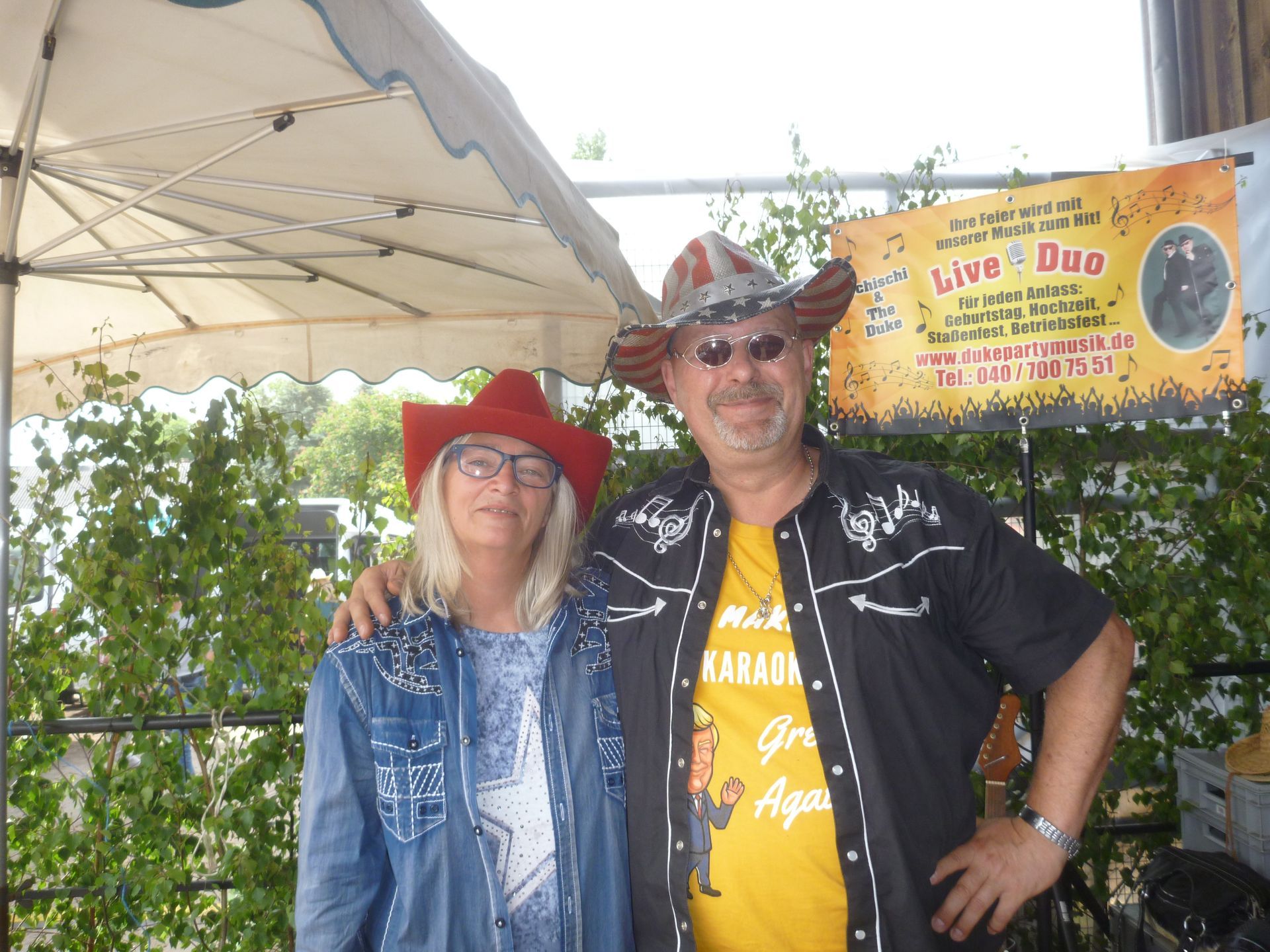 Zwei Personen mit Cowboyhüten posieren unter einem Baldachin. Die eine trägt einen roten Hut und eine Brille, die andere einen gemusterten Hut und ein schwarzes Hemd.