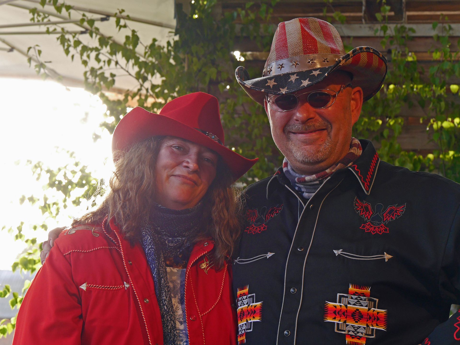Ein Paar in Cowboykleidung, die Frau in Rot, der Mann in Schwarz mit einer Mütze mit amerikanischer Flagge, lächelt draußen.