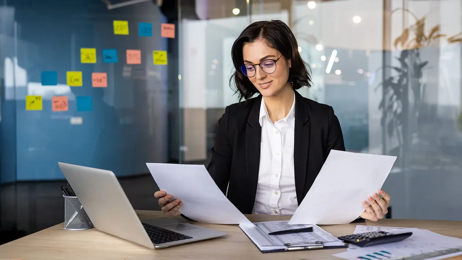Person in glasses reviewing papers beside a laptop in a modern office with colorful sticky notes on glass wall