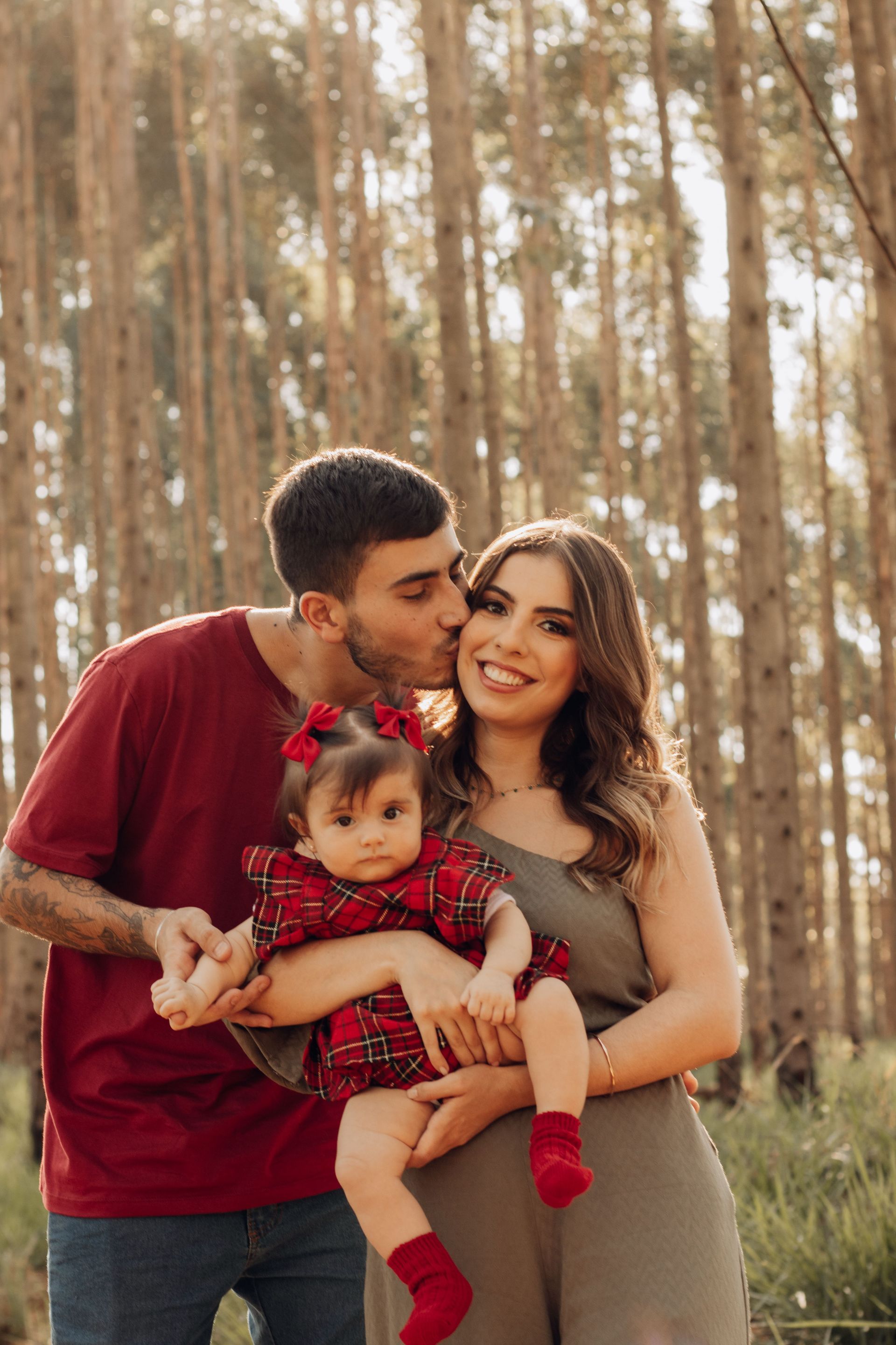 Family of three in a forest, parents holding a toddler; father kisses mother’s cheek.