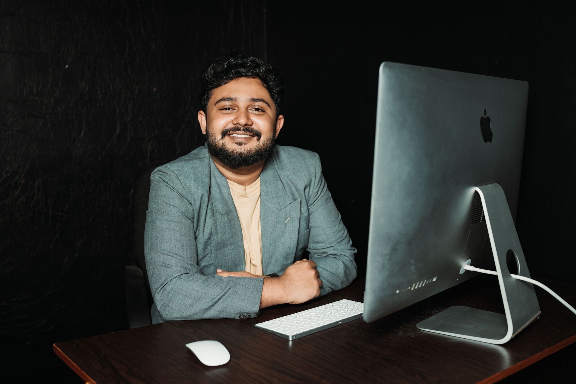 Smiling person at a desk with a large desktop computer and mouse on a dark background
