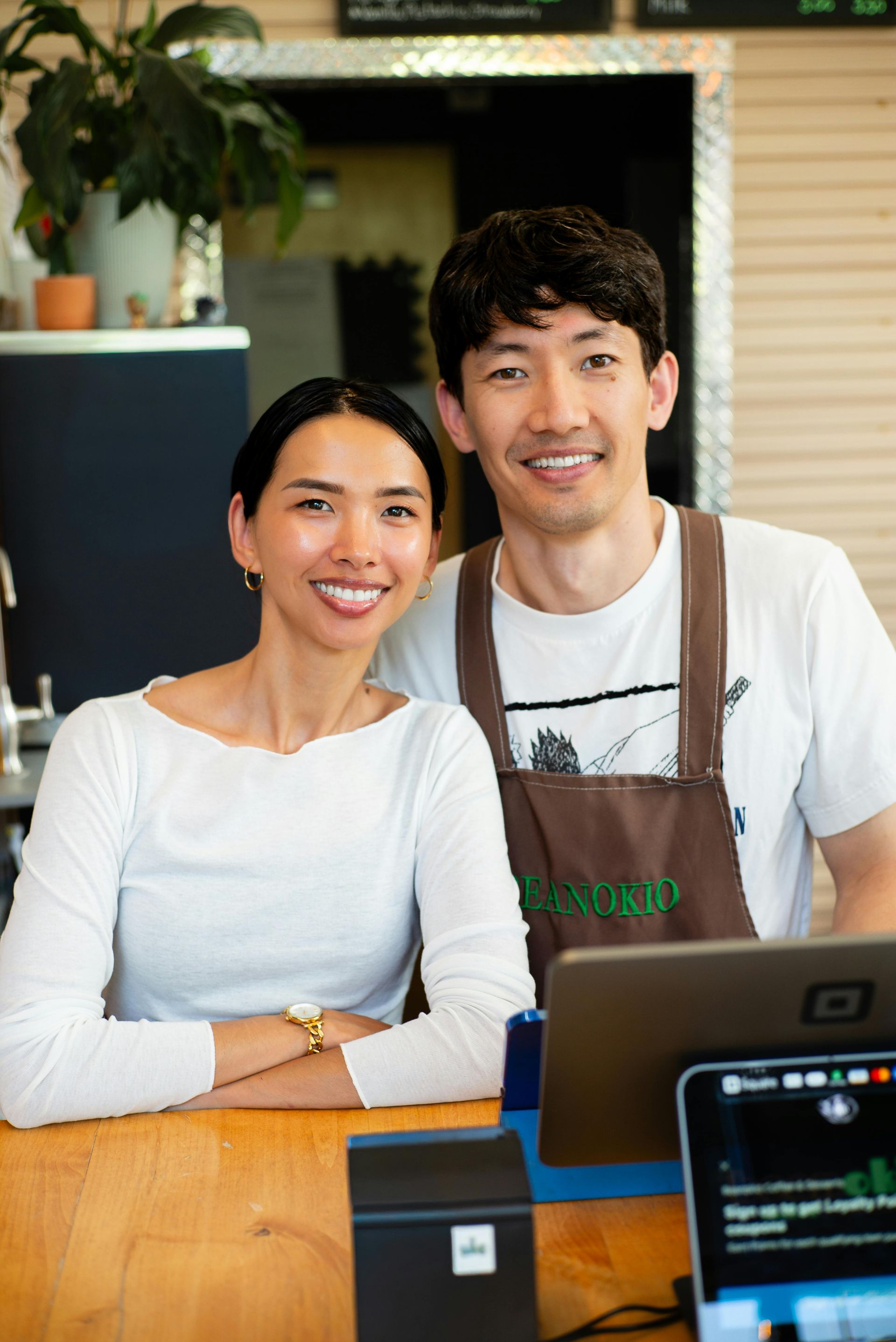 Two smiling café staff behind the counter, with laptop and point-of-sale terminal in front.