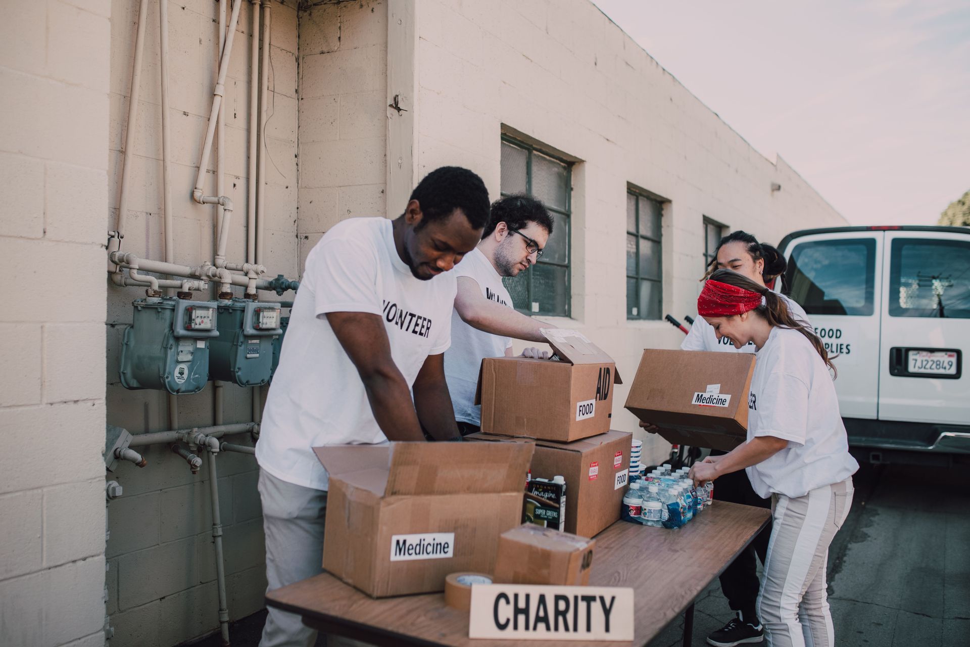 Volunteers packing charity boxes on a table beside a white van outdoors