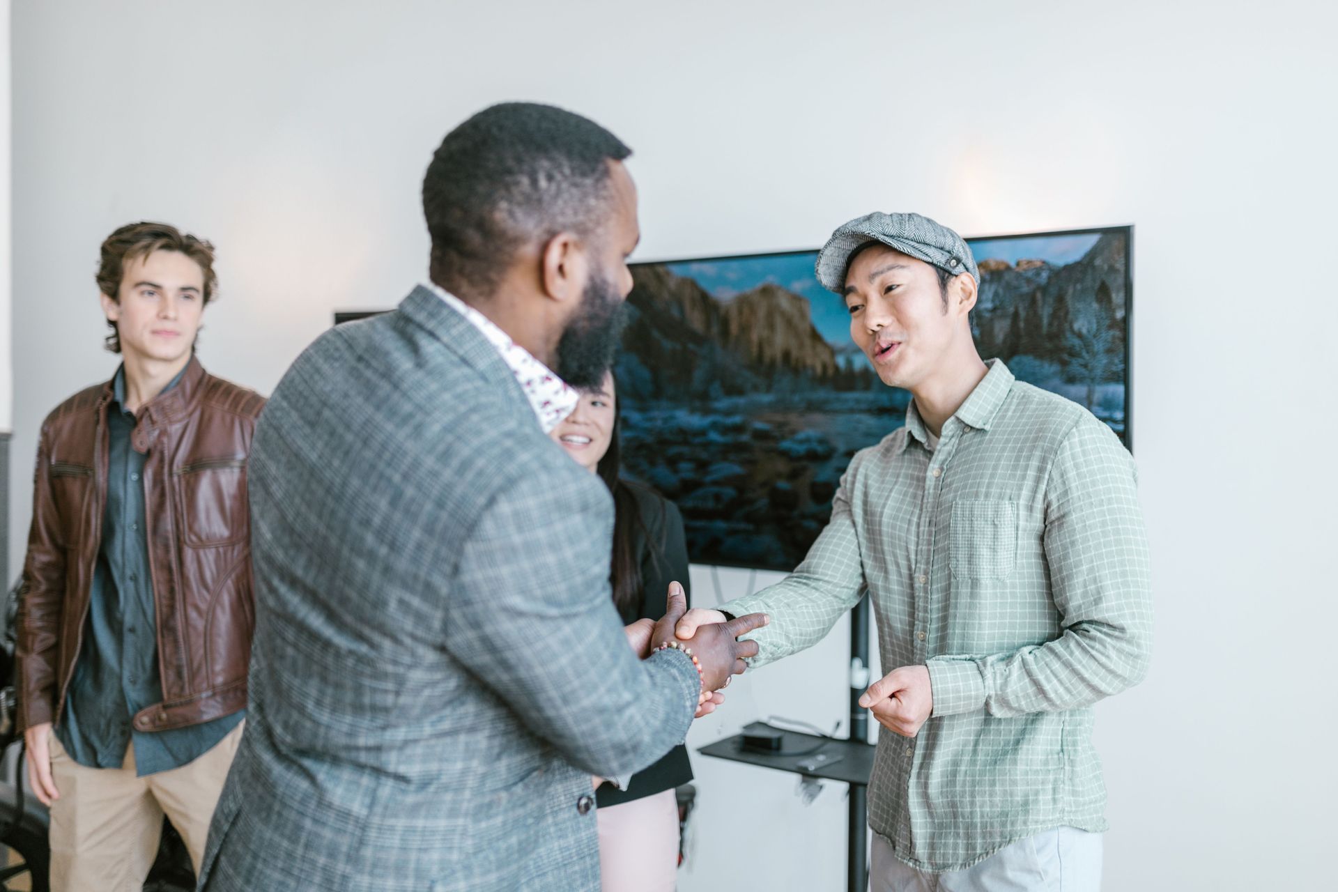 Two people shaking hands in a bright room, with another person standing nearby and a landscape painting in the background.