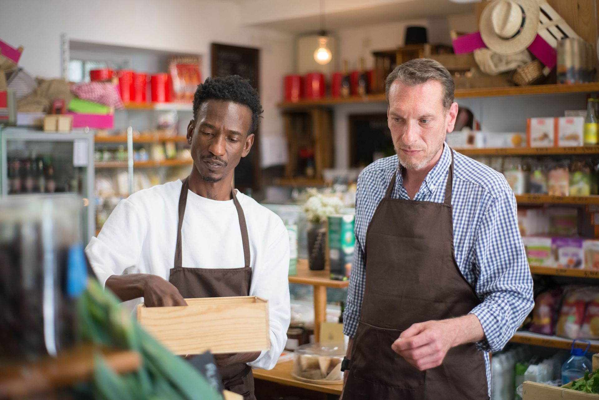 Two men in aprons working in a shop, one holding a wooden crate and one writing.