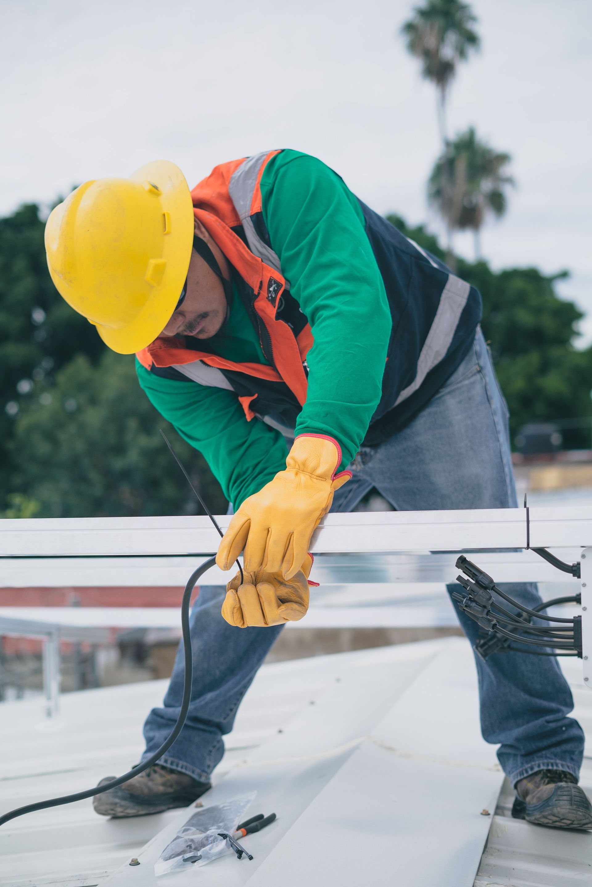 Construction worker in yellow hard hat and gloves bending over a rooftop, inspecting materials.
