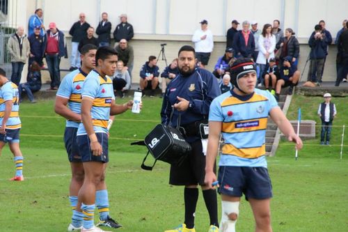 Rugby players in light blue and yellow uniforms stand on a field with a referee and spectators in the background