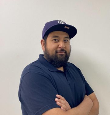 Man in a navy polo and cap stands with arms crossed against a plain wall.