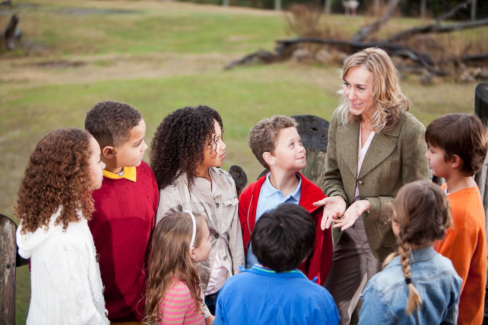 Teacher talking to a group of children outdoors. Smiling, gesturing, near a wooden fence and greenery.