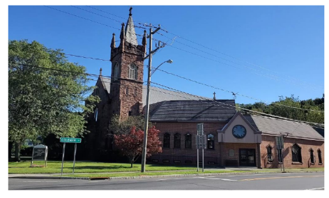 Brick church with tall steeple and arched windows beside a road on a sunny day