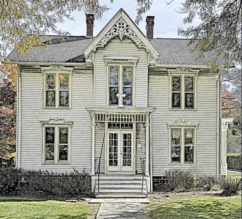 Victorian-style cream house with ornate trim, front steps, and a central double door amidst trees and lawn