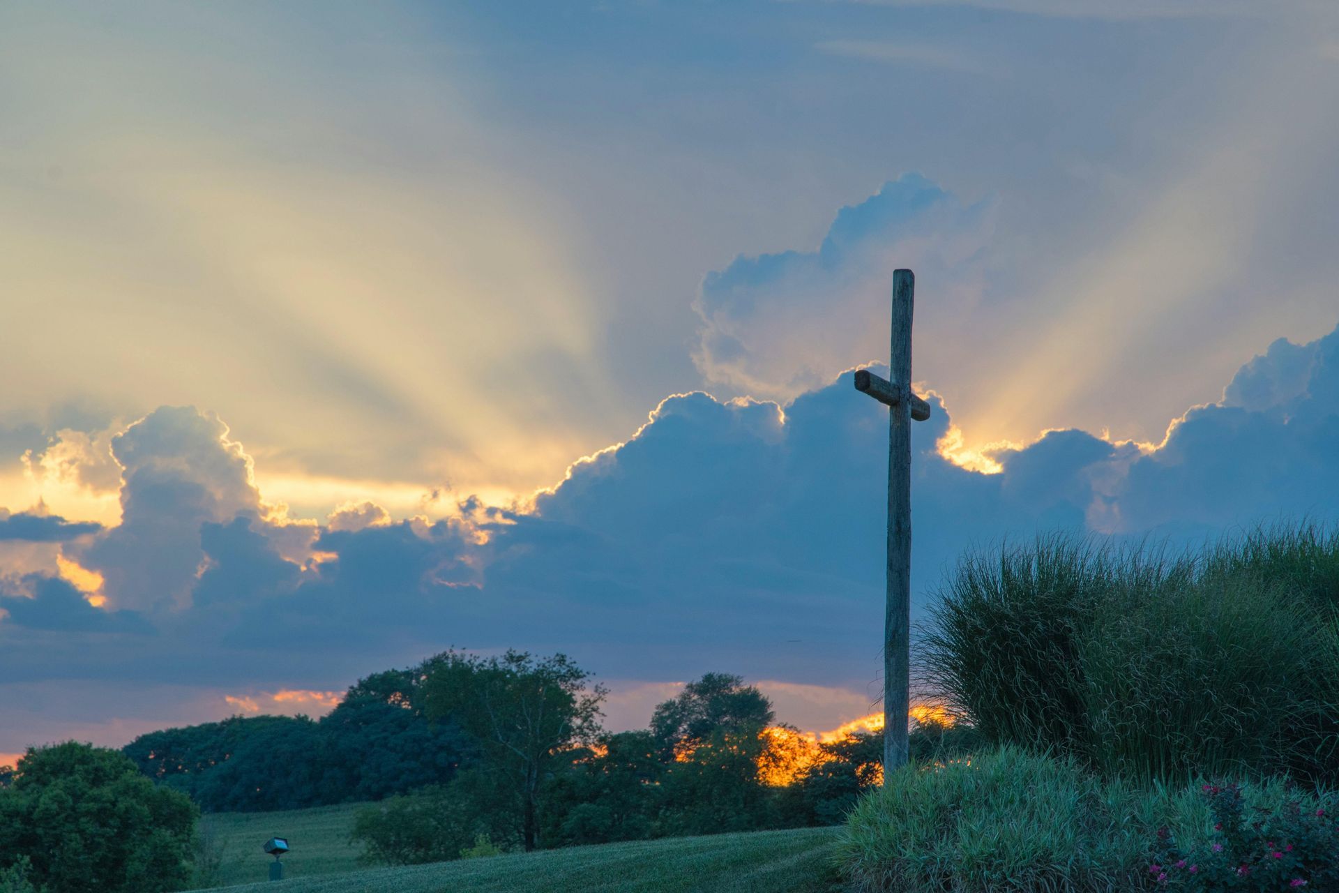 Sun rays behind clouds over a grassy field with a wooden cross at dusk