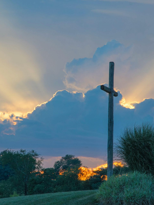 Wooden cross silhouetted against a sunset sky with glowing clouds and trees below