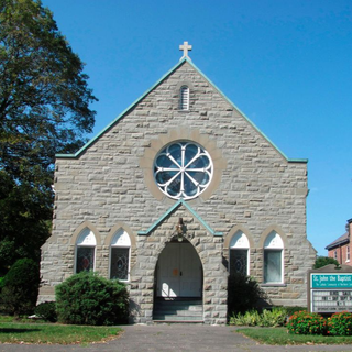 Stone church facade with a cross, rose window, arched doorway, and small sign by the lawn
