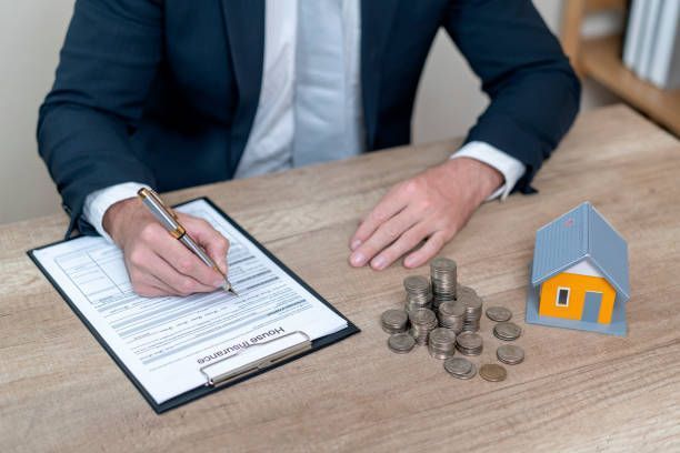 A man is sitting at a desk writing on a clipboard next to a pile of coins and a model house.