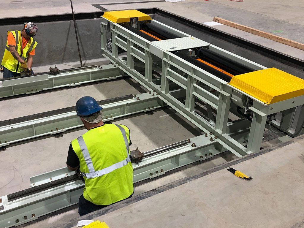 Two workers installing a metal conveyor system, wearing safety vests, in a concrete area.
