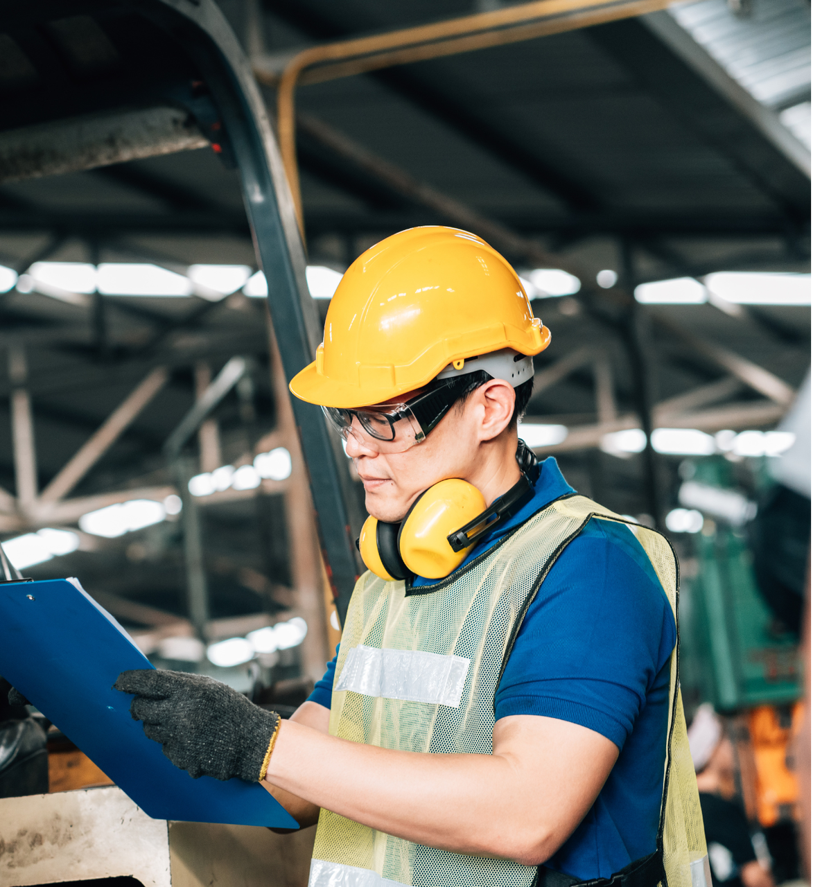 Factory worker in yellow hard hat, safety glasses, and vest, inspecting a clipboard.