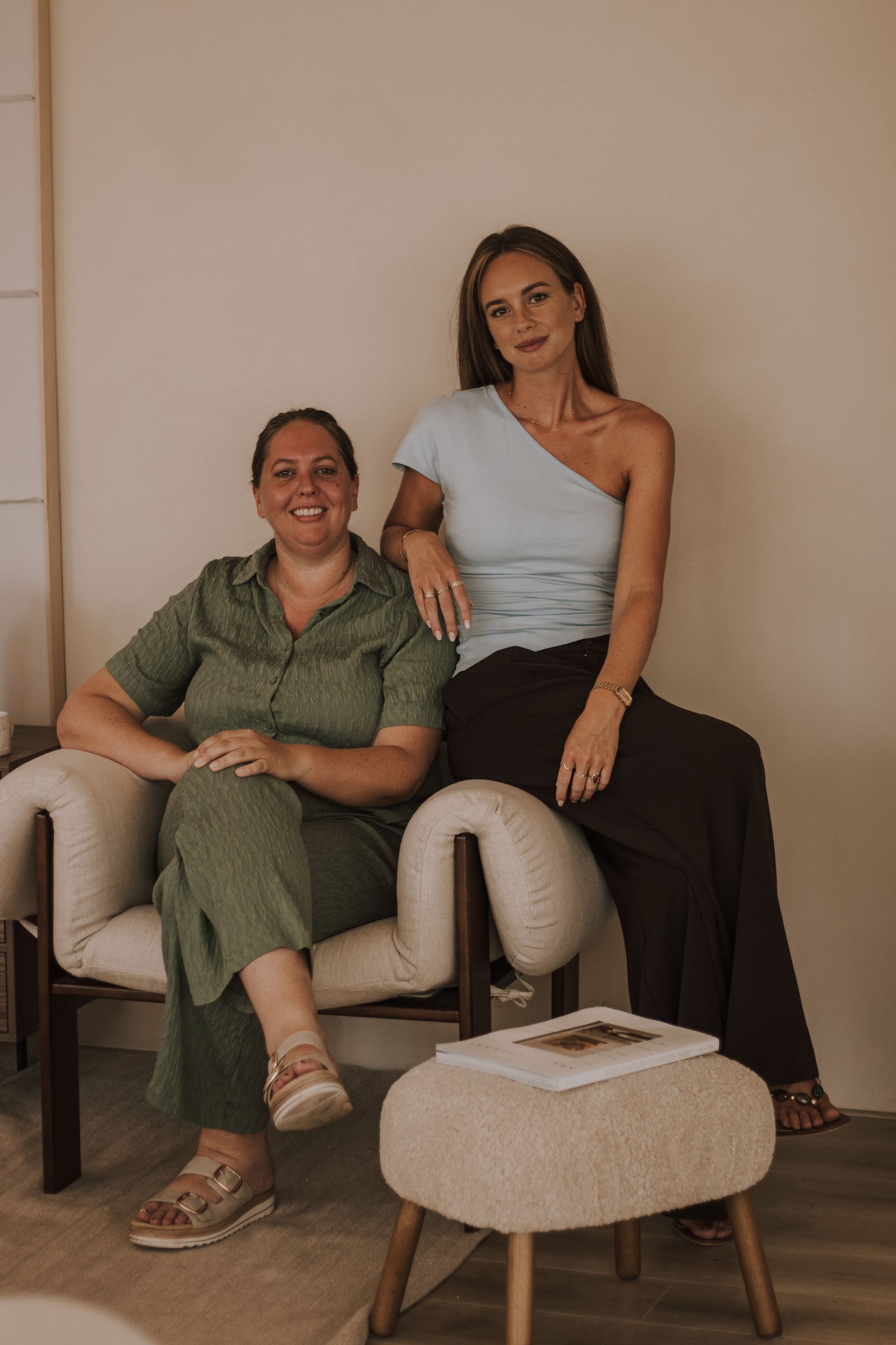 Two women seated in beige chairs in a minimalist living room, one smiling, one in a one-shoulder top.