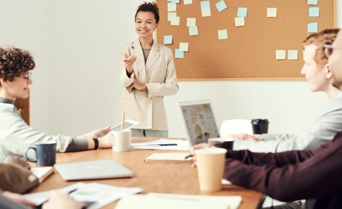 Team meeting in a bright office, with a presenter standing by a corkboard covered in sticky notes.