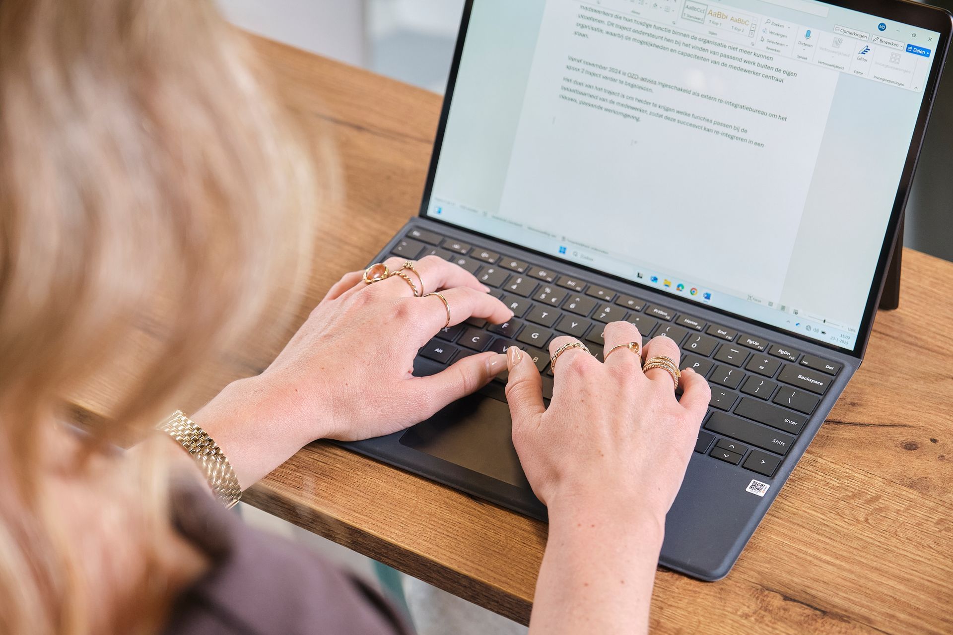 Een persoon typt op een laptop aan een houten tafel, met een document open op het scherm.