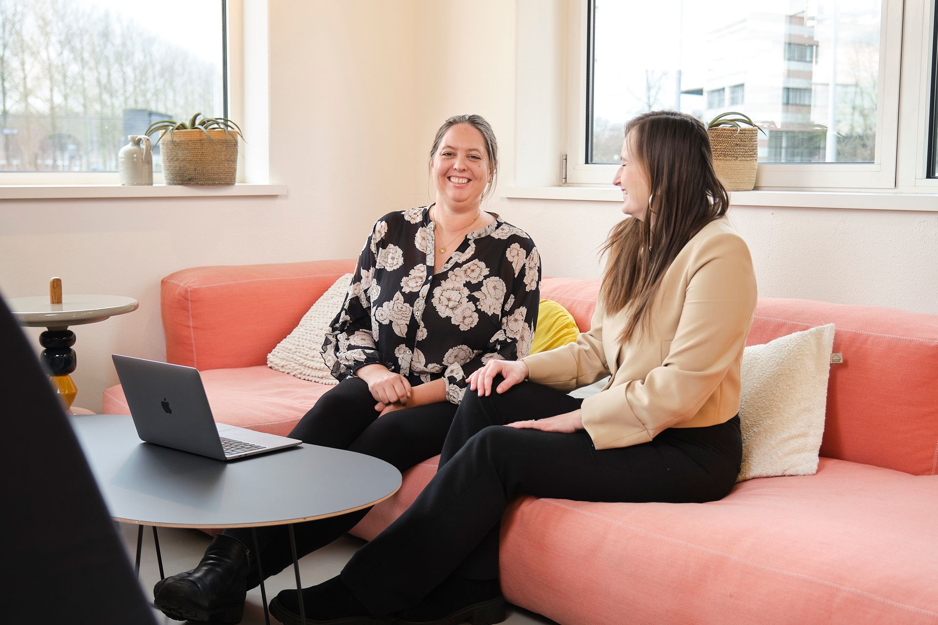 Twee vrouwen praten op een roze bank naast een laptop in een lichte woonkamer.