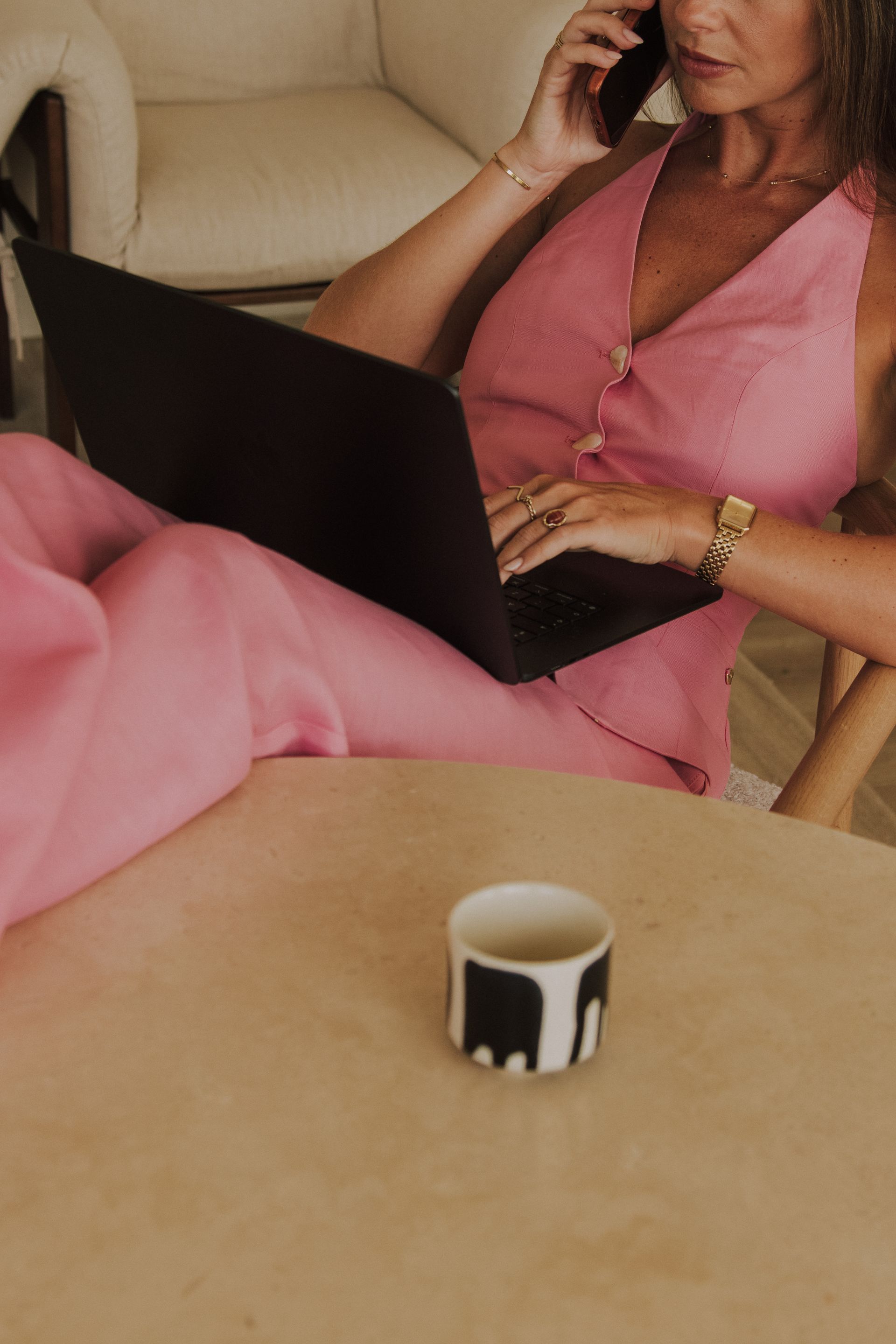 Woman in pink outfit on a phone call beside a laptop at a table with a small mug.
