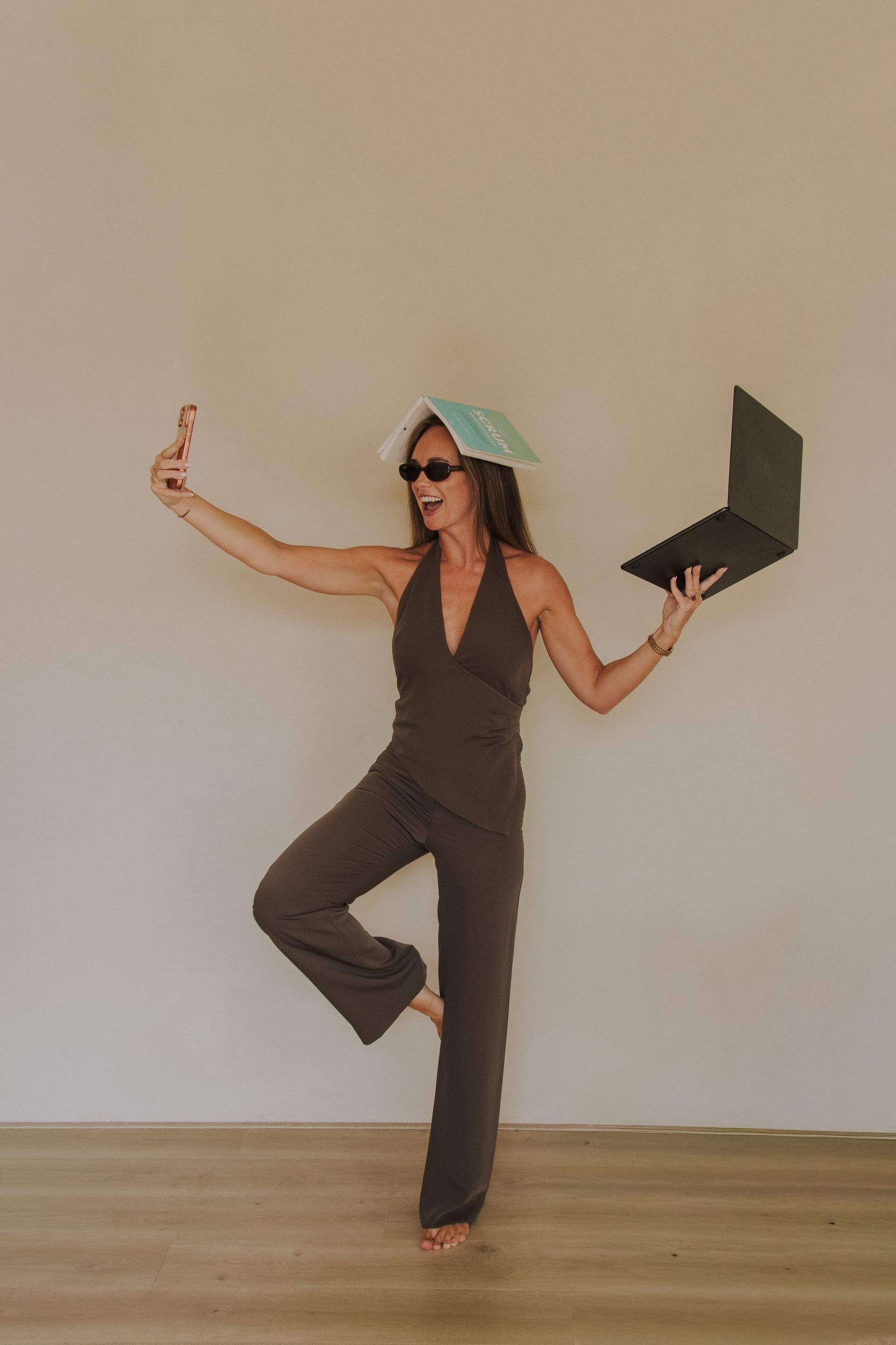 Woman in dark clothing balances on one leg indoors, holding an open black umbrella and a small object.