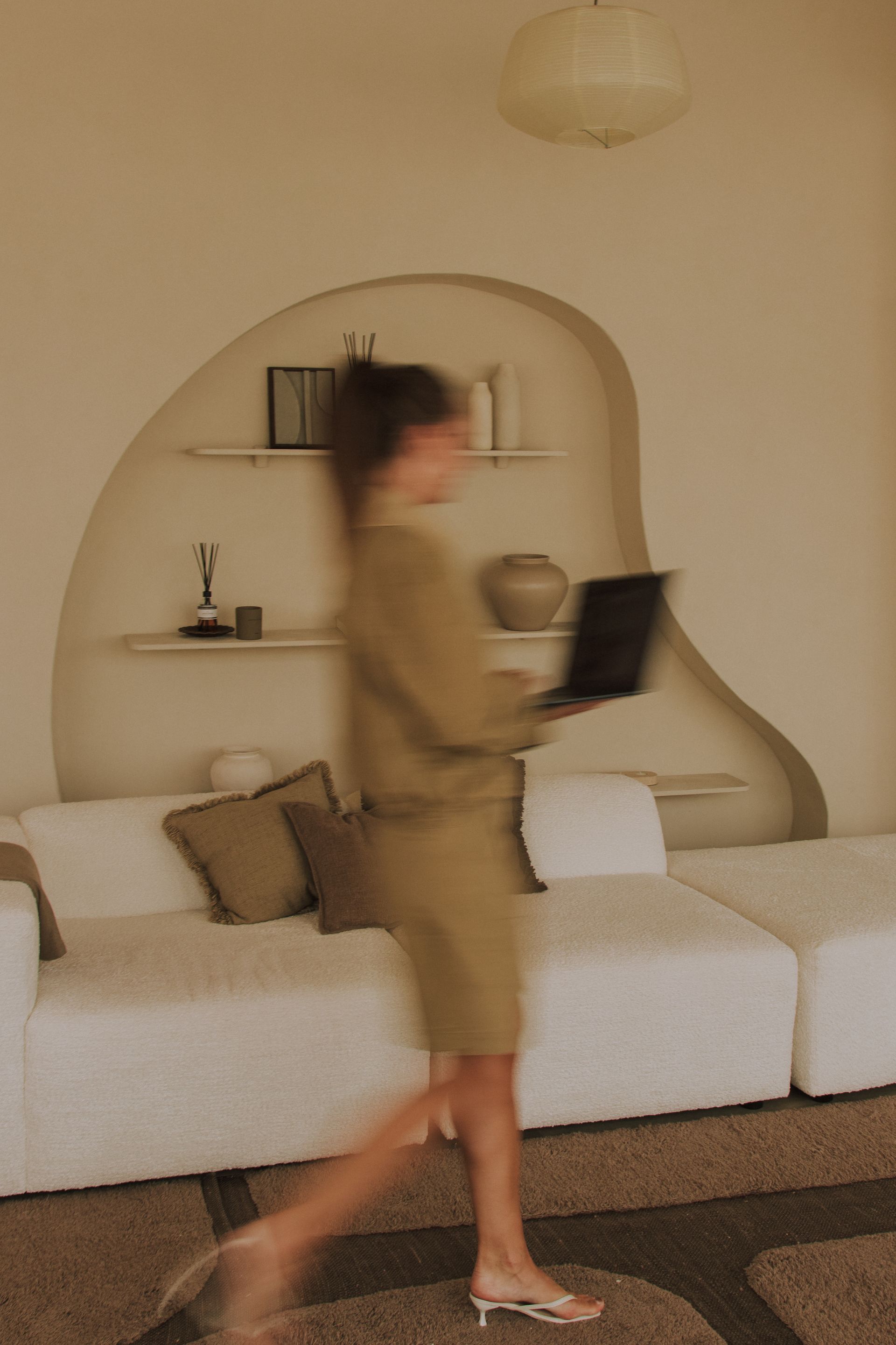 Blurred person walking past a white sofa in a beige living room, holding a laptop.