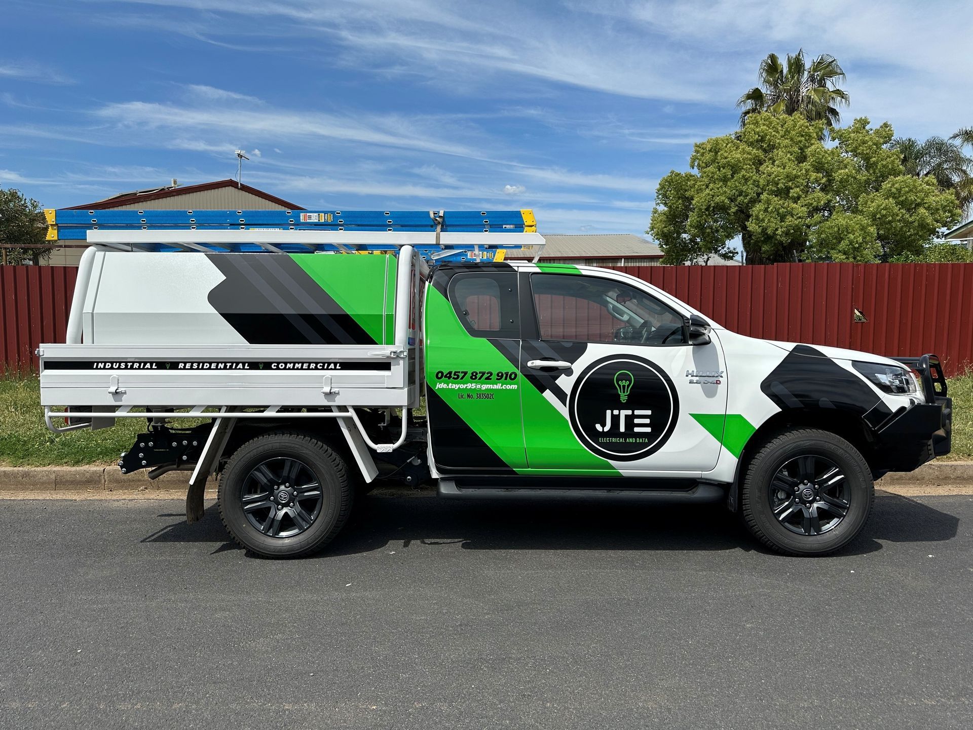 A JTE-branded white, green, and black work truck with a ladder rack parked on a street in front of a red fence.