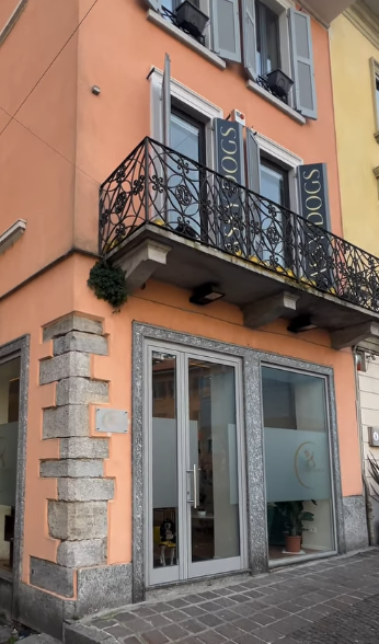 Facciata arancione di un edificio con un balcone in ferro battuto sopra le vetrine dei negozi, affacciata su una strada stretta.