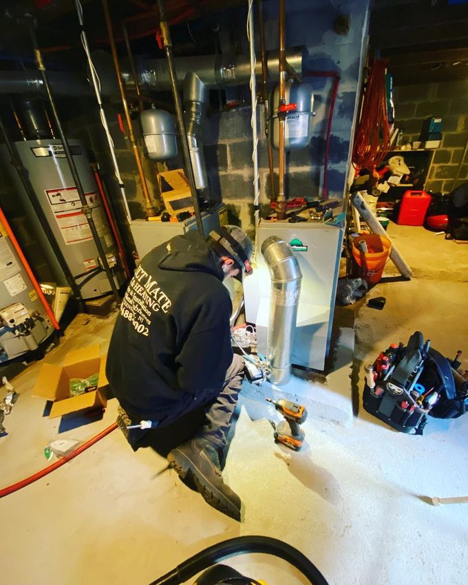 Worker kneeling in a basement utility room, inspecting HVAC equipment and pipes near exposed ducts.