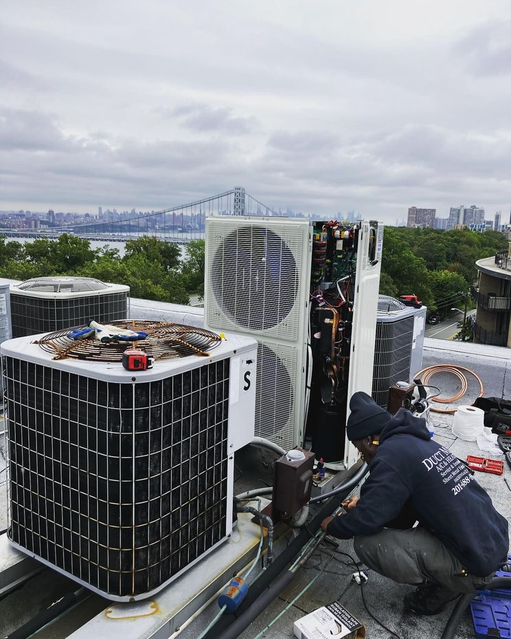 Technicians repairing rooftop HVAC units with a city skyline and bridge in the background
