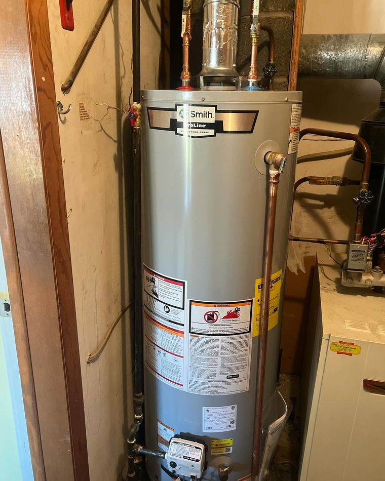 Tall silver water heater in a basement utility area with pipes and labels attached