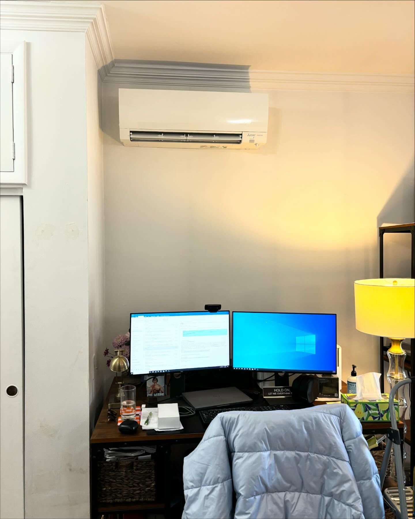 Home office desk with dual monitors, chair, lamp, and wall-mounted air conditioner.