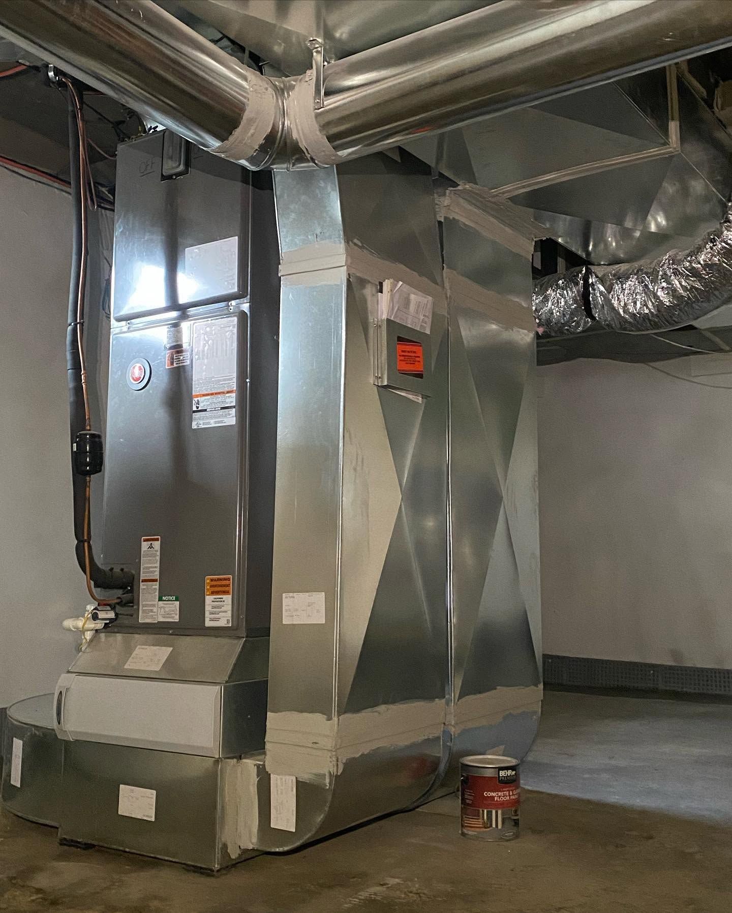 Large metallic HVAC duct system in a mechanical room with exposed pipes and ceiling insulation.